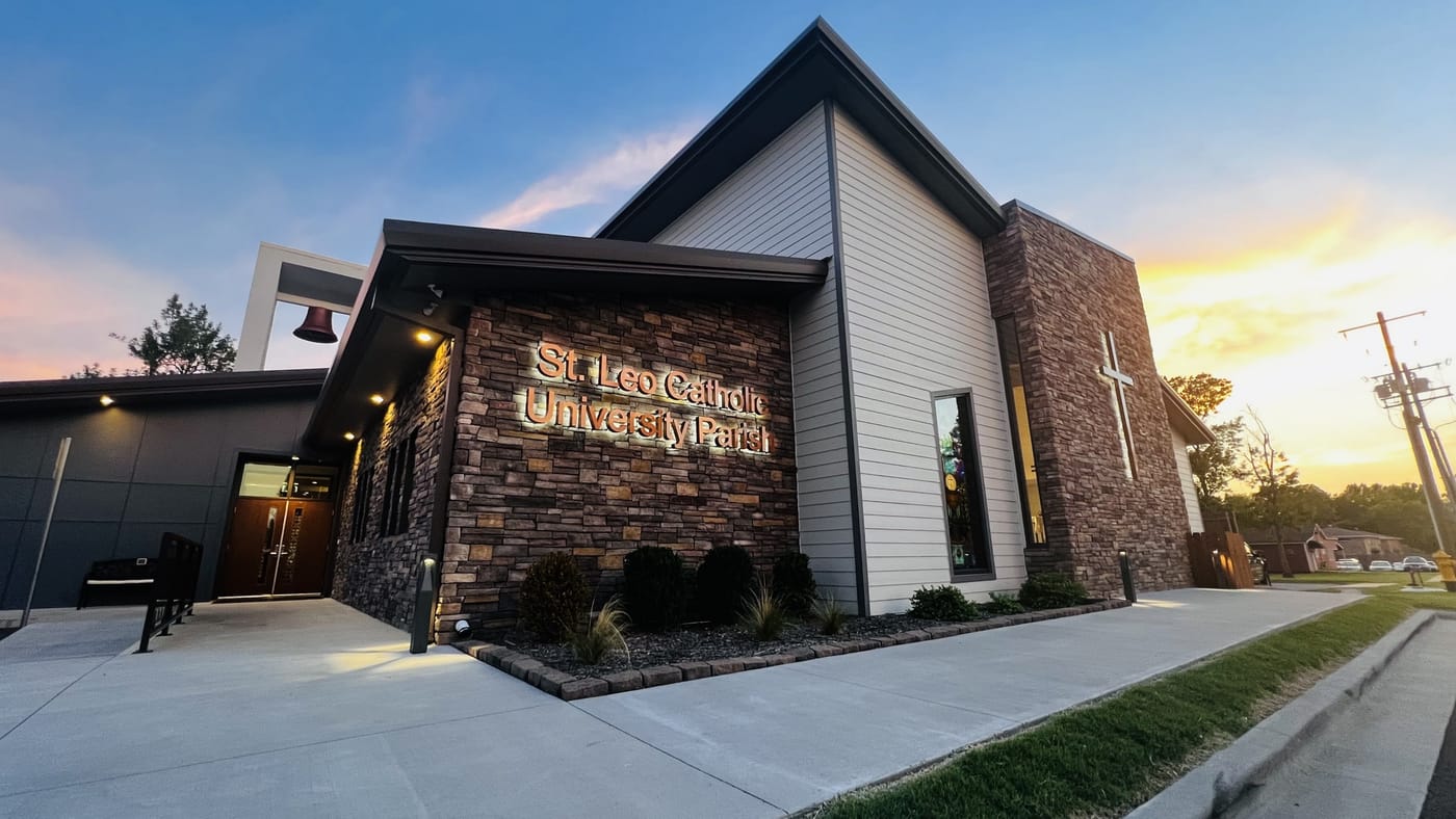St. Leo Catholic University Parish at sunset — the stone front with the lit parish sign, the bell tower, and the cross.